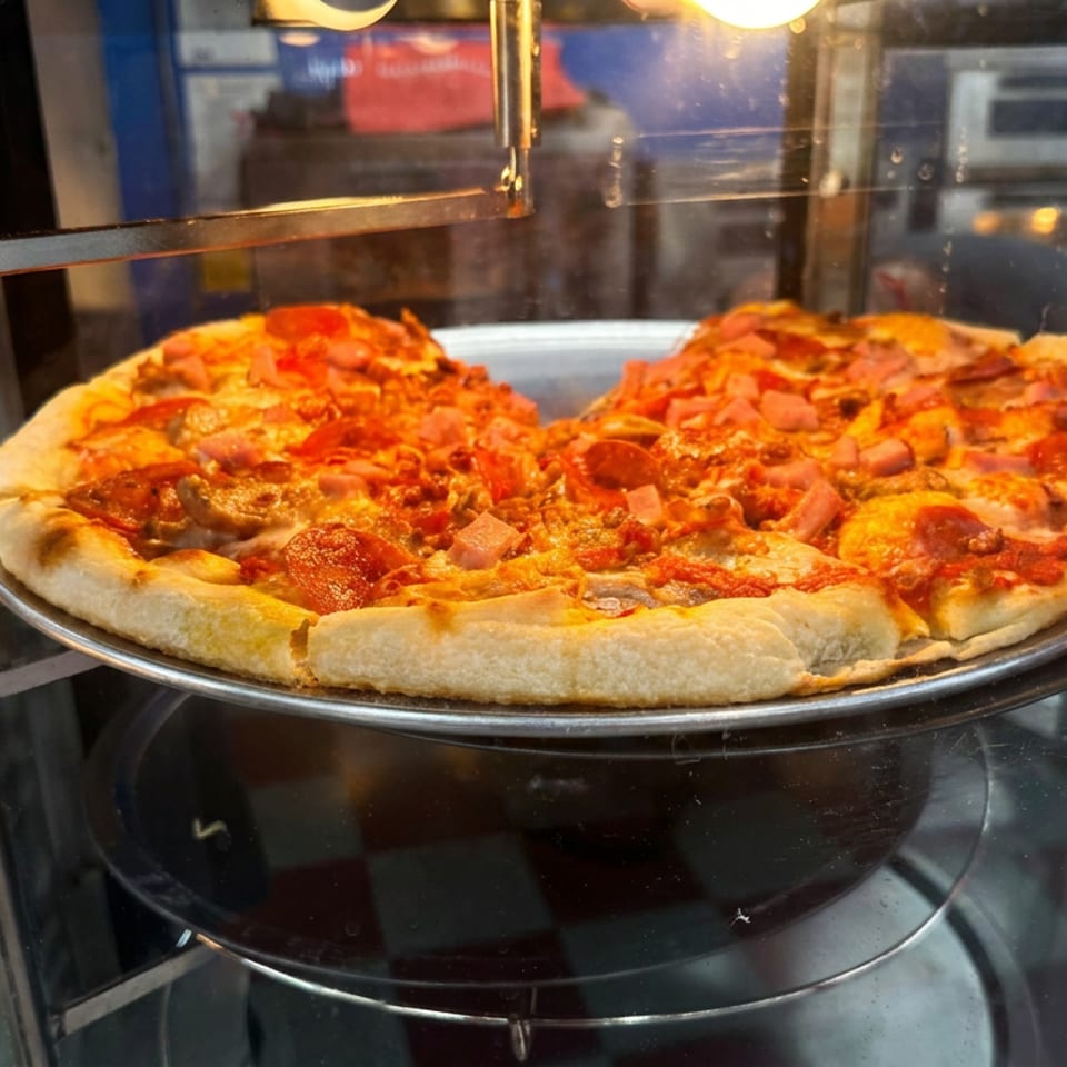 Pepperoni and ham pizza with melted cheese displayed on a metal tray inside a glass warming case.
